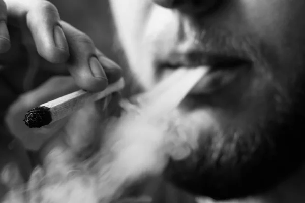 A man smokes cannabis weed, a joint and a lighter in his hands. Smoke on a black background. Concepts of medical marijuana use and legalization of the cannabis.Black and white — Stock Photo, Image