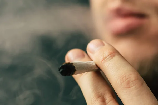 Un hombre fuma marihuana, un porro y un encendedor en sus manos. Humo sobre fondo negro. Conceptos de uso médico de la marihuana y legalización del cannabis . — Foto de Stock