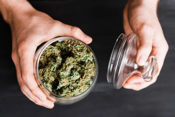 Ovanifrån. En burk med knoppar av cannabis i händerna på en man. Begreppet laglig marijuana är medicinsk ogräs. På en svart bakgrund. — Stockfoto