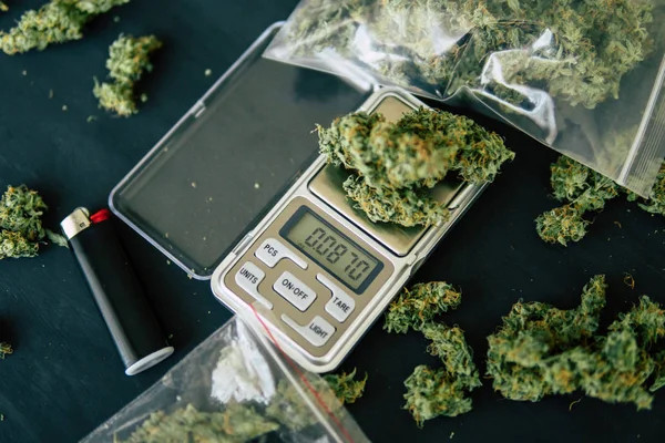 Конусы цветов марихуаны на весах, шлифовальный и измельченный косяк конопли и пакет травы и смартфона на фоне черного дерева — стоковое фото