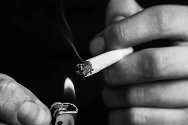 Un homme fume de l'herbe de cannabis, un joint et un briquet dans ses mains. Fumée sur fond noir. Concepts de la consommation de marijuana médicale et de la légalisation du cannabis.Noir et blanc — Photo