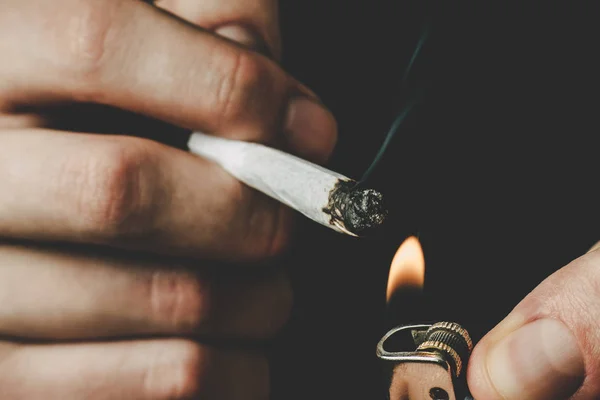 Cannabis weed A man smokes, a joint and a lighter in his hands. Smoke on a black background. Concepts of medical marijuana use and legalization of the cannabis. — Stock Photo, Image