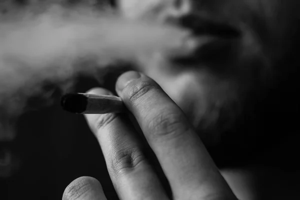 Un homme fume de l'herbe de cannabis, un joint et un briquet dans ses mains. Fumée sur fond noir. Concepts de la consommation de marijuana médicale et de la légalisation du cannabis.Noir et blanc — Photo