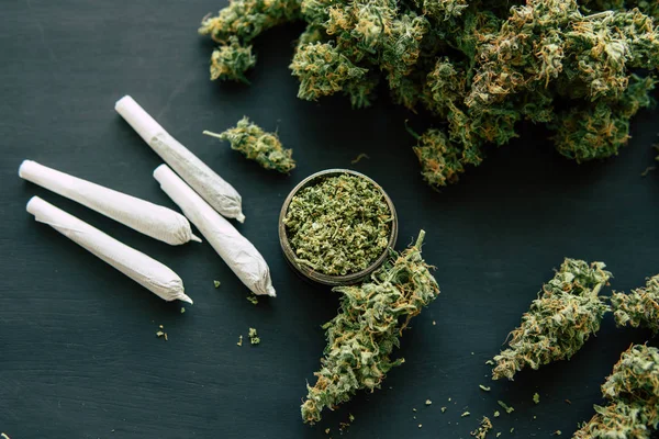 Joint and Grinder with crushed cannabis flowers and and weed on the background of a black wooden table top view — Stock Photo, Image