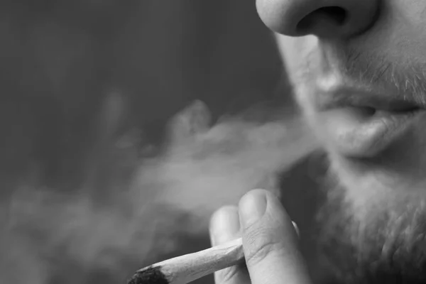 Un homme fume de l'herbe de cannabis, un joint et un briquet dans ses mains. Fumée sur fond noir. Concepts de la consommation de marijuana médicale et de la légalisation du cannabis.Noir et blanc — Photo