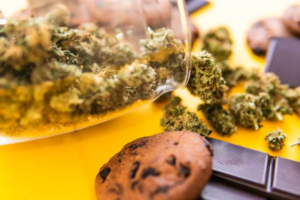 Treatment of medical marijuana for use in food, yellow background. Cannabis CBD herb Chocolate and Cookies. Cookies and Chocolate with weed and buds of marijuana on the table.