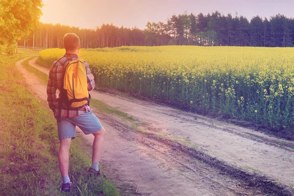 Turista está andando ao longo da estrada caminho com uma mochila perto do prado ao pôr do sol — Fotografia de Stock