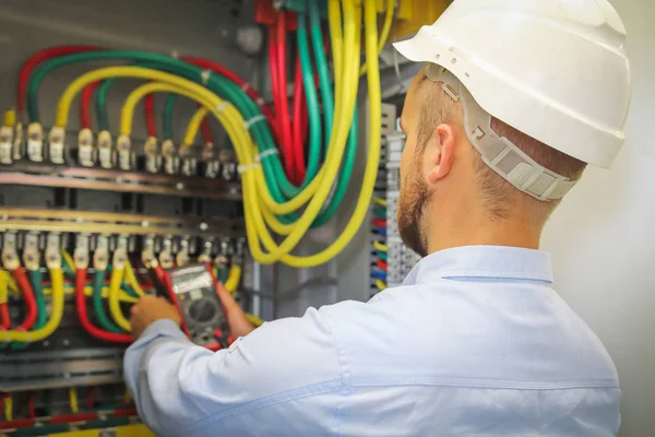 Engenheiro eletricista mede a tensão dos cabos de energia elétrica no gabinete. trabalhador em capacete branco com multímetro — Fotografia de Stock