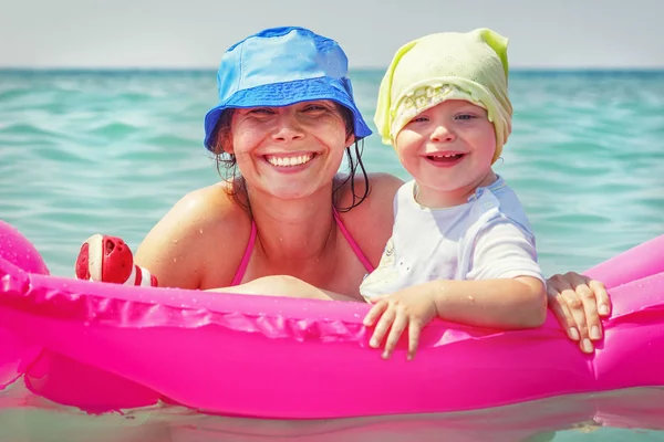 Mamá y su hija están nadando en agua de mar azul en colchón inflable. Feliz familia sonriente de vacaciones. Mujer y niño bañándose en un día soleado brillante en el mar . — Foto de Stock