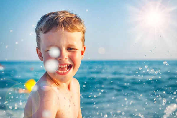 Niño feliz baña y salpica en agua de mar en día soleado brillante. Niño caucásico hacer Salpicaduras y gotas de agua. Vacaciones de verano en el mar. Niño en la playa. Concepto de vacaciones — Foto de Stock