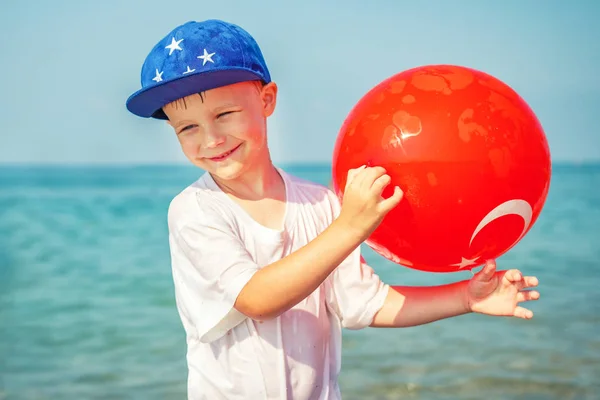 Smiling European boy at sea with ball of Turkish flag. Anak bahagia dengan latar belakang air laut biru. Liburan musim panas di Turki — Stok Foto