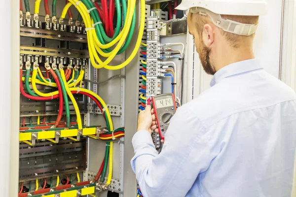 Elektriker ingenjör i vit hjälm testar power spänning panel av trefas krets. Serviceteknikern elektrisk med multimeter. — Stockfoto