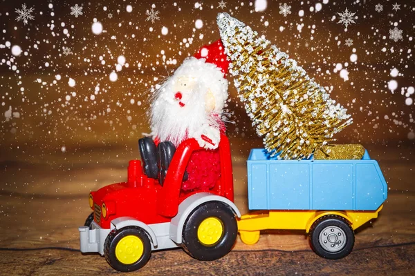 Christmas holiday celebration theme. Santa Claus carries Christmas tree on tractor with trailer. Xmas background. New Year concept. Winter snowflakes on wooden background. Snowfall on X-mas toys.
