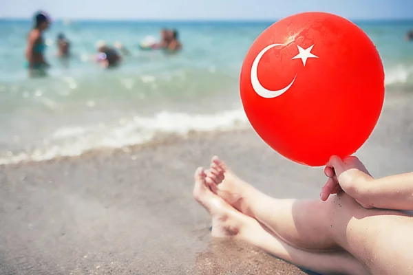 Young woman relaxes on tropical sea beach and holds red balloon with turkish flag on it. Summer vacation on sunny day at sea beach in Turkey. Legs of girl on sea on background of bathing people. — Stock Photo, Image