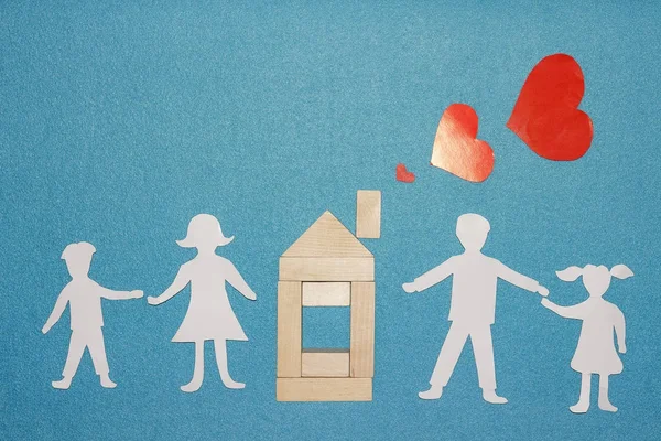 Amore in casa e concetto di famiglia. Famiglia di carta che si tiene per mano e casa in legno con cuori rossi dal tubo del camino su sfondo blu — Foto Stock