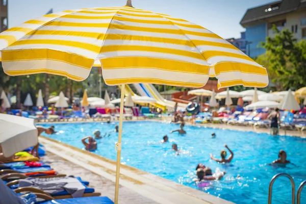 People bathe in pool on sunny summer day. Sunny yellow umbrella on background of pool with blue water. Beach vacation. Concept of summer holidays.