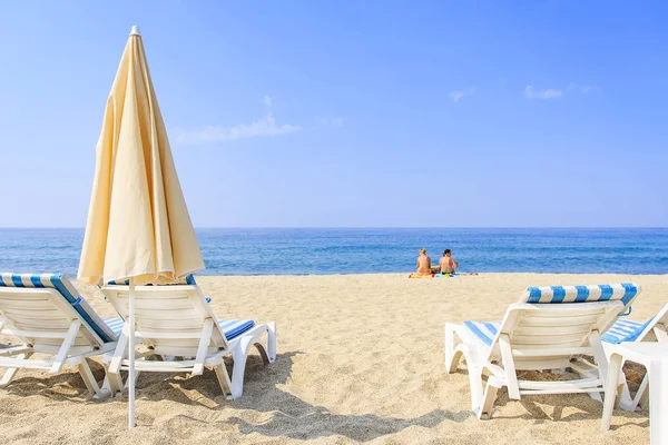 Zonnebaden op resort strand op warme, zonnige dag. Parasols en ligbedden op witte zand tegen de blauwe zee en de heldere hemel. Twee vrouwen op zand zitten en ontspannen in de buurt van de zee. — Stockfoto