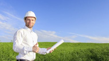 Mühendis Oluşturucu beyaz kask ve yeşil alan arka planda parlak güneşli bir günde üzerinde planları ile. Mühendislik çözümleri tasarım ve inşaat için. Başarılı çalışan.