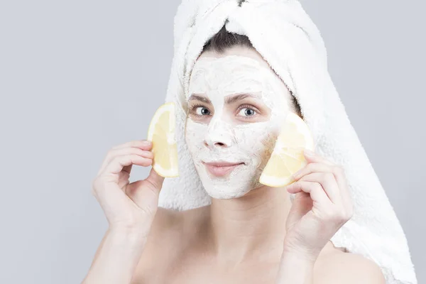 Wanita muda cantik menyaksikan masker krem wajah memegang dua potong lemon. Konsep perawatan kulit. Gadis dengan krim pelembab masker di wajah dan handuk di kepala . — Stok Foto