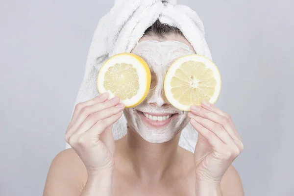 Senyum brunette wanita memegang dua potong lemon di depan wajahnya. Wanita dengan masker wajah lembab. Konsep kecantikan dan perawatan kulit . — Stok Foto
