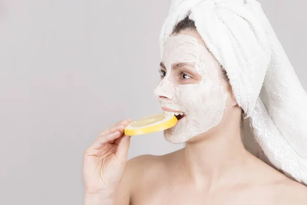 Koncepcja opieka piękna i skóry. Młoda kobieta jedzenie plasterkiem cytryny. Dziewczyna z nawilżająca maseczka na twarz. Dziewczyna z ręcznikiem na głowie i krem maska na twarz. — Zdjęcie stockowe