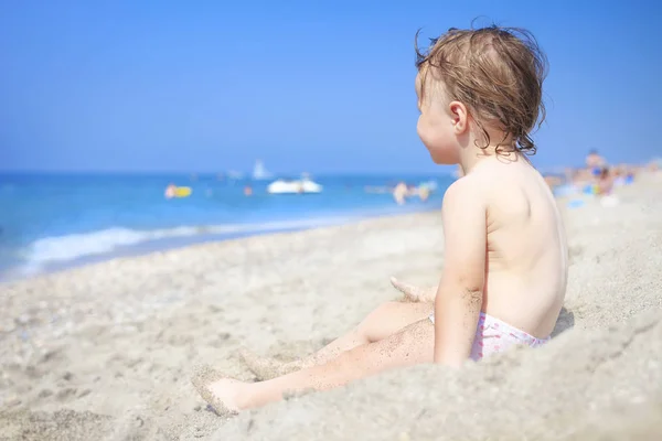 Niño pequeño en la playa de arena de mar mirando las olas. Niño en el mar en claro día soleado. Relájese en la playa con niños. Niño en la arena blanca en la playa . — Foto de Stock