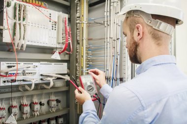 Hizmet adam elinde multimetre ile elektrik kontrol paneli ayarlar. Mühendis kask içinde koruma aygıtı sınar. İşçi elektrik Endüstri/sanayii donatımı denetler. Mühendislik çalışmaları otomasyon kutusunda.