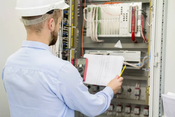 Ingeniero en casco blanco lee dibujo de diseño contra panel industrial eléctrico. Trabajador de servicio analiza el circuito eléctrico en la caja de control de automatización eléctrica con controladores . — Foto de Stock