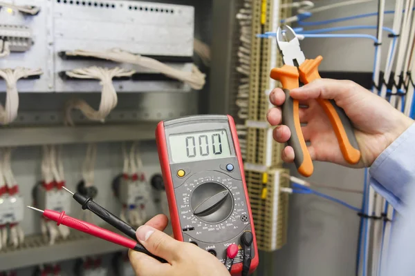 Testador e cortadores de fio em mãos de eletricista contra painel de controle elétrico de equipamentos de automação. ferramentas de engenharia na mão do engenheiro para ajustar controladores elétricos — Fotografia de Stock