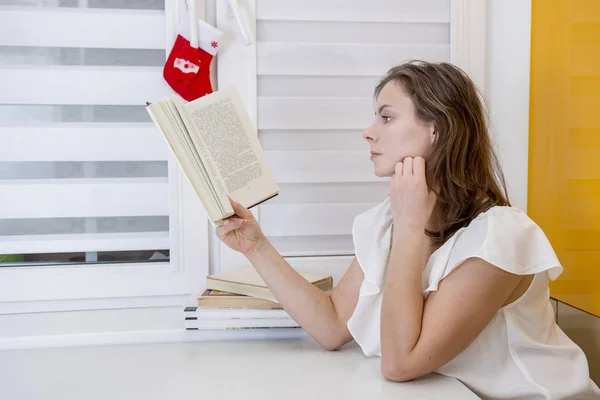 Giovane donna attraente si siede al tavolo e legge libro a casa — Foto Stock