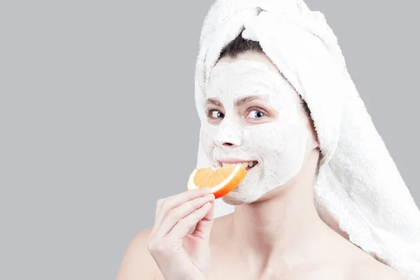 Wanita muda yang menarik dengan masker wajah menggigit jeruk dan tersenyum. Kecantikan, spa, perawatan kulit dan tubuh — Stok Foto