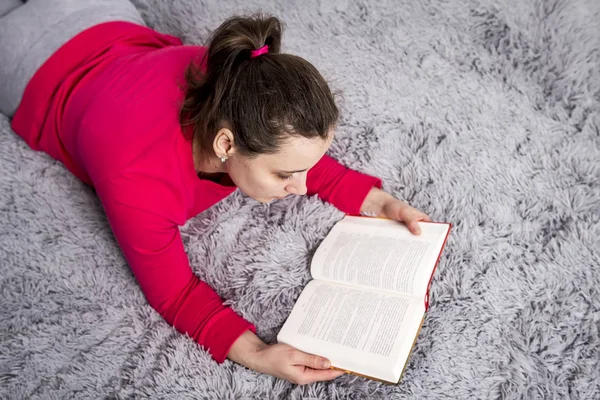 Vacker brunett ung kvinna läser bok på sängen i mysig lägenhet. Hobby läsa böcker och fiktion. Spendera tid med boken. En tjej läser skönlitteratur hemma. — Stockfoto