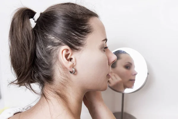 Beautiful young woman in front of round mirror. Attractive girl doing makeup at home. Caucasian woman looks after the skin of the face in front of the mirror.