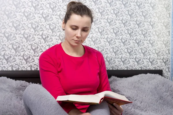 Una giovane donna legge un libro a casa. Una ragazza è seduta sul letto a leggere un romanzo. Fantasia dell'hobby. Trascorrere del tempo con il libro — Foto Stock