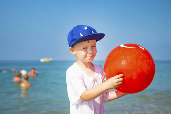 Retrato de niño en la playa. Niño en gorra con una pelota inflable contra el mar azul en un día de verano claro y soleado de vacaciones. Feliz niño sonriendo en el mar. Jugar a los niños en la playa . — Foto de Stock