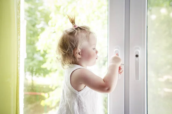 Small child is standing on windowsill and opens window. Locks on windows prevent children from falling out of window. Girl playing with window handle. — Stock Photo, Image