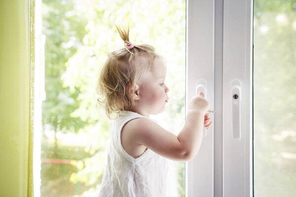 small child is standing on windowsill and opens window. Locks on windows prevent children from falling out of window. Girl playing with window handle.