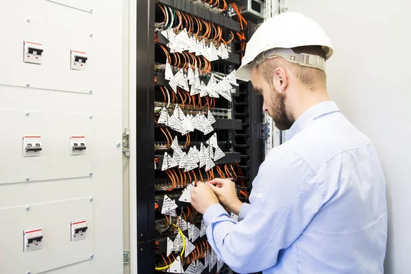 Engenheiro executa ajuste de gabinete de rede de computador com controladores, interruptores e cabos ópticos. Um trabalhador em capacete branco se conecta — Fotografia de Stock