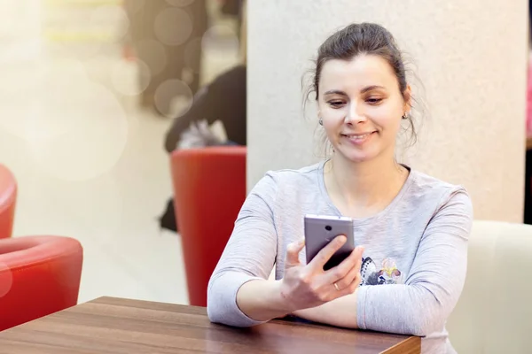 Young brunette woman in cafe uses smartphone and smiles. The girl is in the wi fi wi-fi zone of the cafe. Free internet in the restaurant — Stock Photo, Image