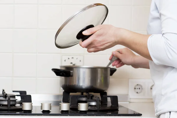 Woman is cooking soup in kitchen. housewife prepares food at home. caucasian woman holds lid from saucepan in her hand and stirs soup in saucepan. — Stock Photo, Image