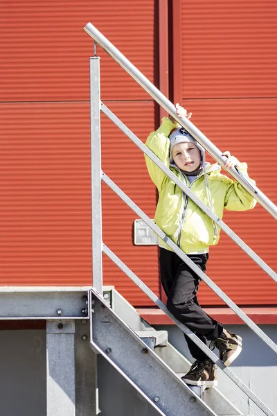 Potret anak laki-laki Kaukasia kecil di topi berpegangan pada tangga rel terhadap dinding logam merah di halaman kota . — Stok Foto