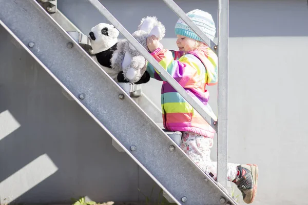 Gadis kecil bermain di tangga logam dengan mainan mewah. Bayi perempuan berjaket berwarna dan topi bermain di dekat rumah . — Stok Foto