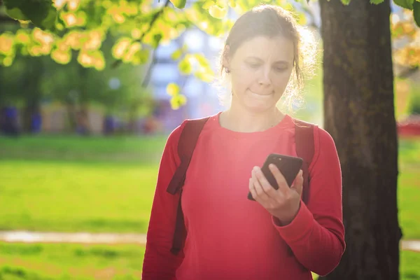 Pensive girl with smartphone in hands in city park. woman is looking at the screen of mobile phone against background of green trees. girl reads news from smartphone. — Stock Photo, Image