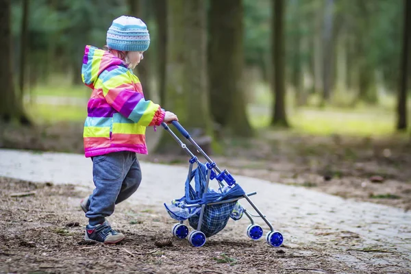 Seorang gadis kecil mainan kereta bayi bergulir di taman. Anak di taman bermain dengan kereta bayi . — Stok Foto