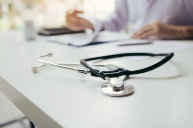 Doktor, elinde kalem tutarak hastanın geçmişini not defterine yazıyor ve hastayla ilaç ve tedavi hakkında konuşuyor..