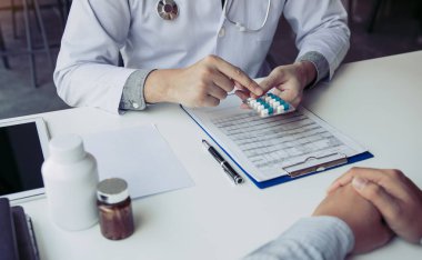 Kendine güveni tam bir doktor elinde bir ilaç şişesi, kıdemli bir hastayla konuşurken yazı yazıyor ve muayenehanede ilaçlarını gözden geçiriyor..