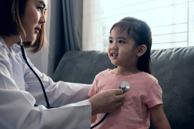 Kadın doktor kalp atışlarını dinlemek için steteskop kullanırken Asyalı kız çocuk gülümsüyor ve tıbbi muayene sırasında koltukta oturuyor.. 