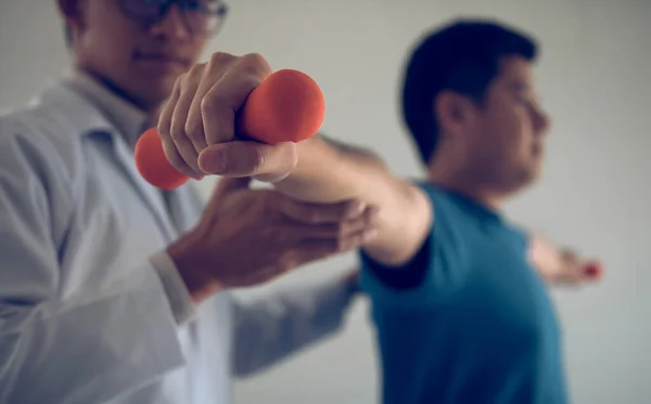 Asian physiotherapist helping a patient lifting dumbbells work through his recovery with weights in clinic room.