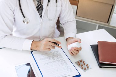 Kendine güveni tam bir doktor elinde bir ilaç şişesi, kıdemli bir hastayla konuşurken yazı yazıyor ve muayenehanede ilaçlarını gözden geçiriyor..