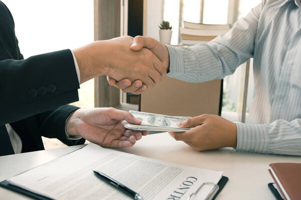 Два корпоративных бизнесмена пожимают друг другу руки, в то время как один человек кладет деньги на документ в офисе с коррупционной концепцией.